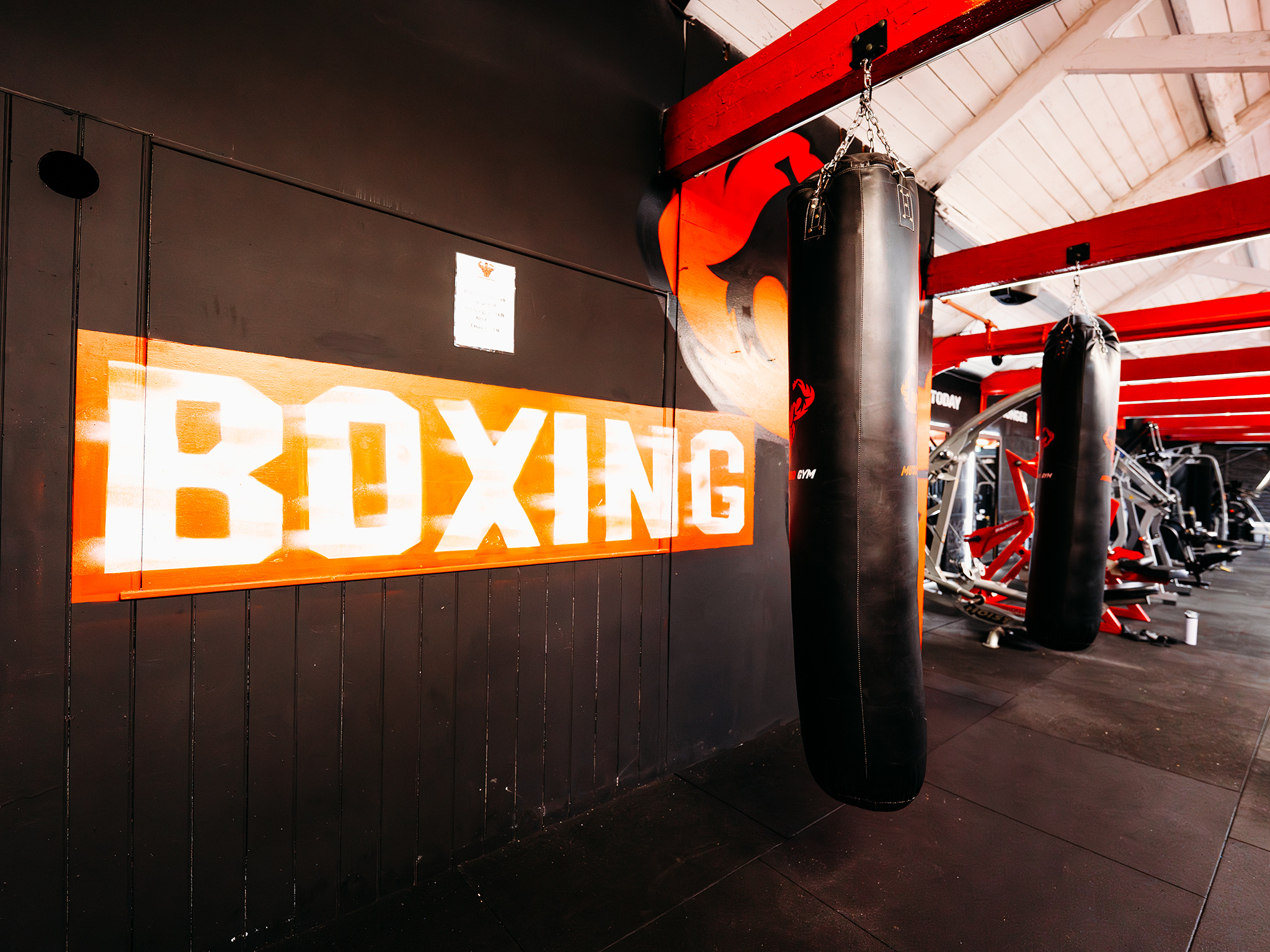 Two black punching bags hang from red ceiling beams in a modern gym’s boxing area. A bold orange wall sign reading “BOXING” sits behind them, with weight machines and benches visible in the warmly lit background.