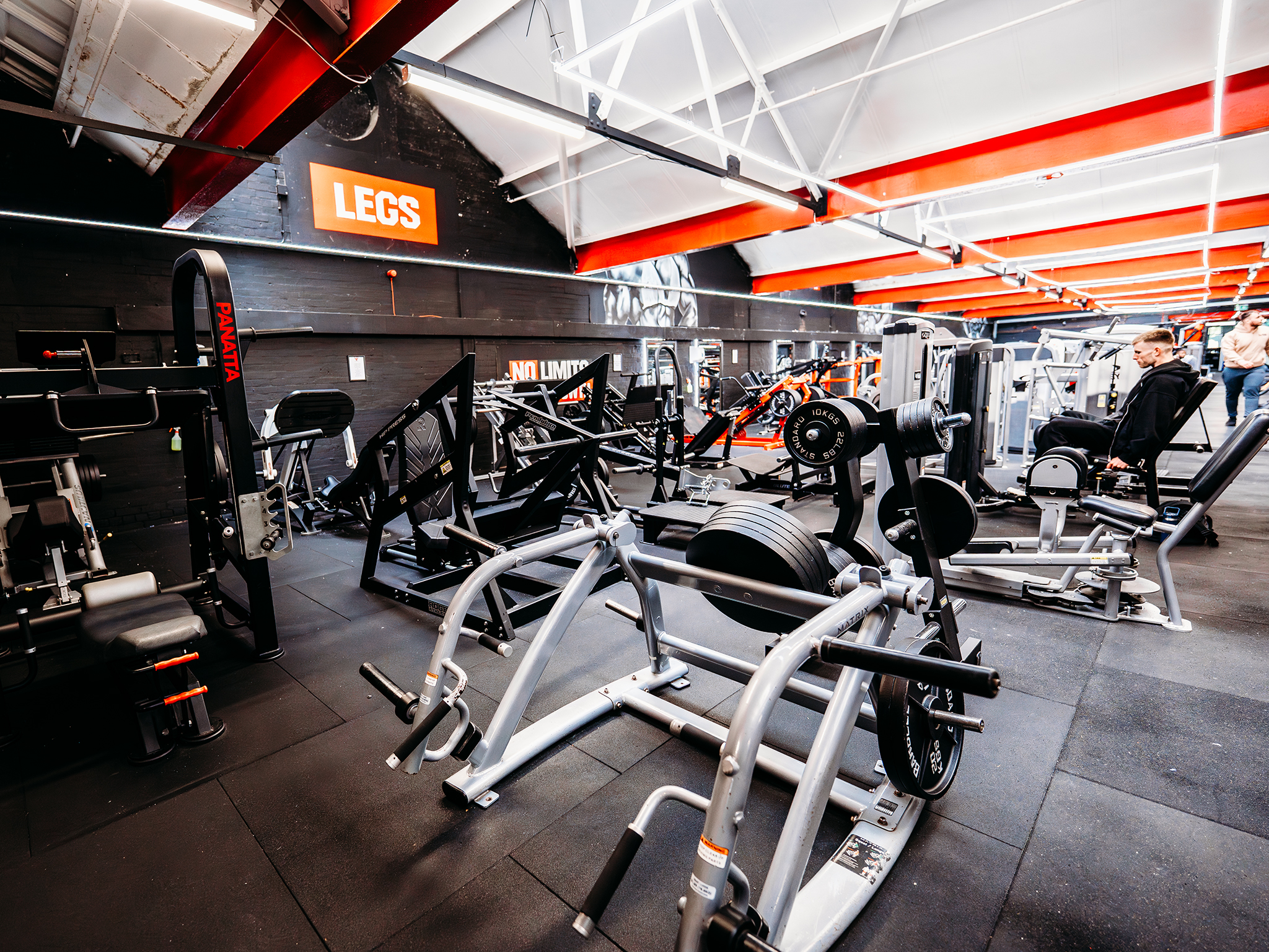 A dedicated leg‑training area in a gym with multiple machines, including leg presses, hack squats, and leg extensions. Black flooring and red‑and‑white ceiling beams frame the space, with an orange “LEGS” sign on the wall and a person using one of the machines.