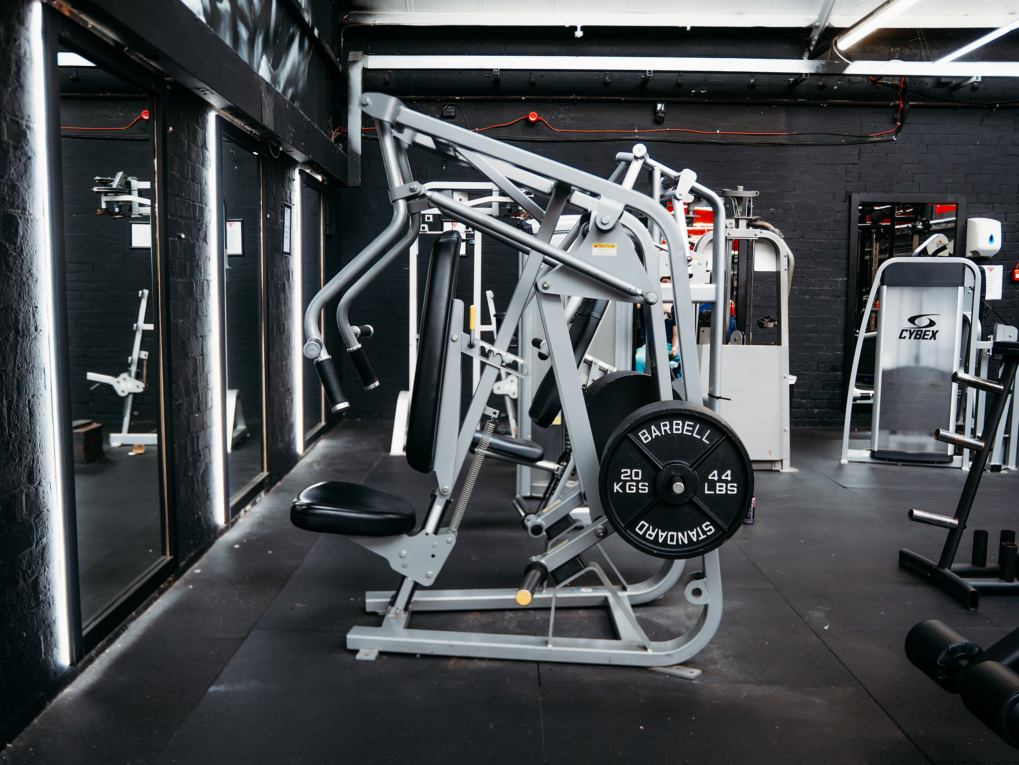 A seated chest‑press machine loaded with multiple 20‑kilogram weight plates sits in a modern gym. The machine has padded seats and handles, with other equipment, mirrors, and black brick walls illuminated by vertical lights in the background.