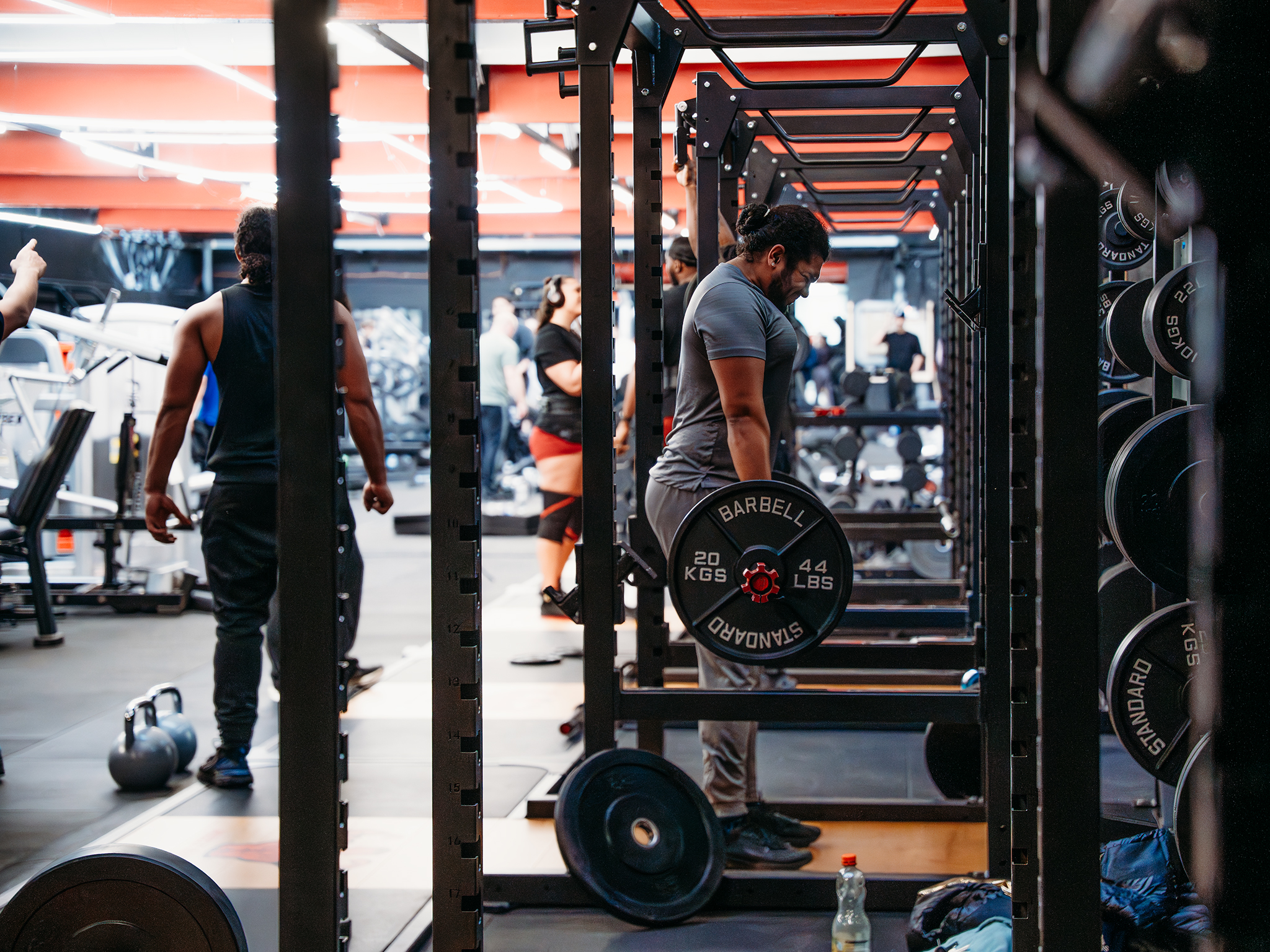 A person performing a deadlift inside a squat rack in a busy gym, lifting a barbell loaded with 20‑kilogram plates while other people train in the background.