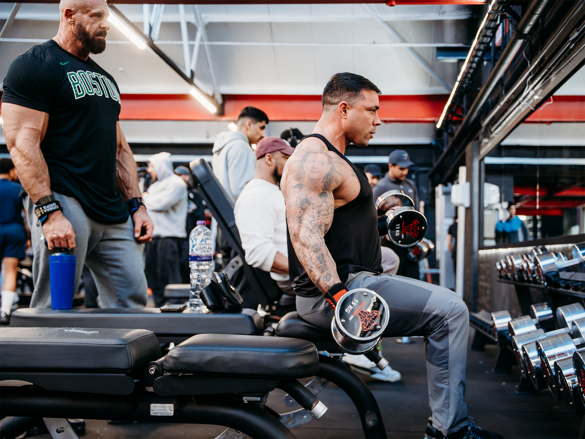 A tattooed person seated on a gym bench performing dumbbell curls with two 15‑kilogram dumbbells, while another person stands nearby holding a shaker bottle and others work out in the background near rows of dumbbells and mirrors