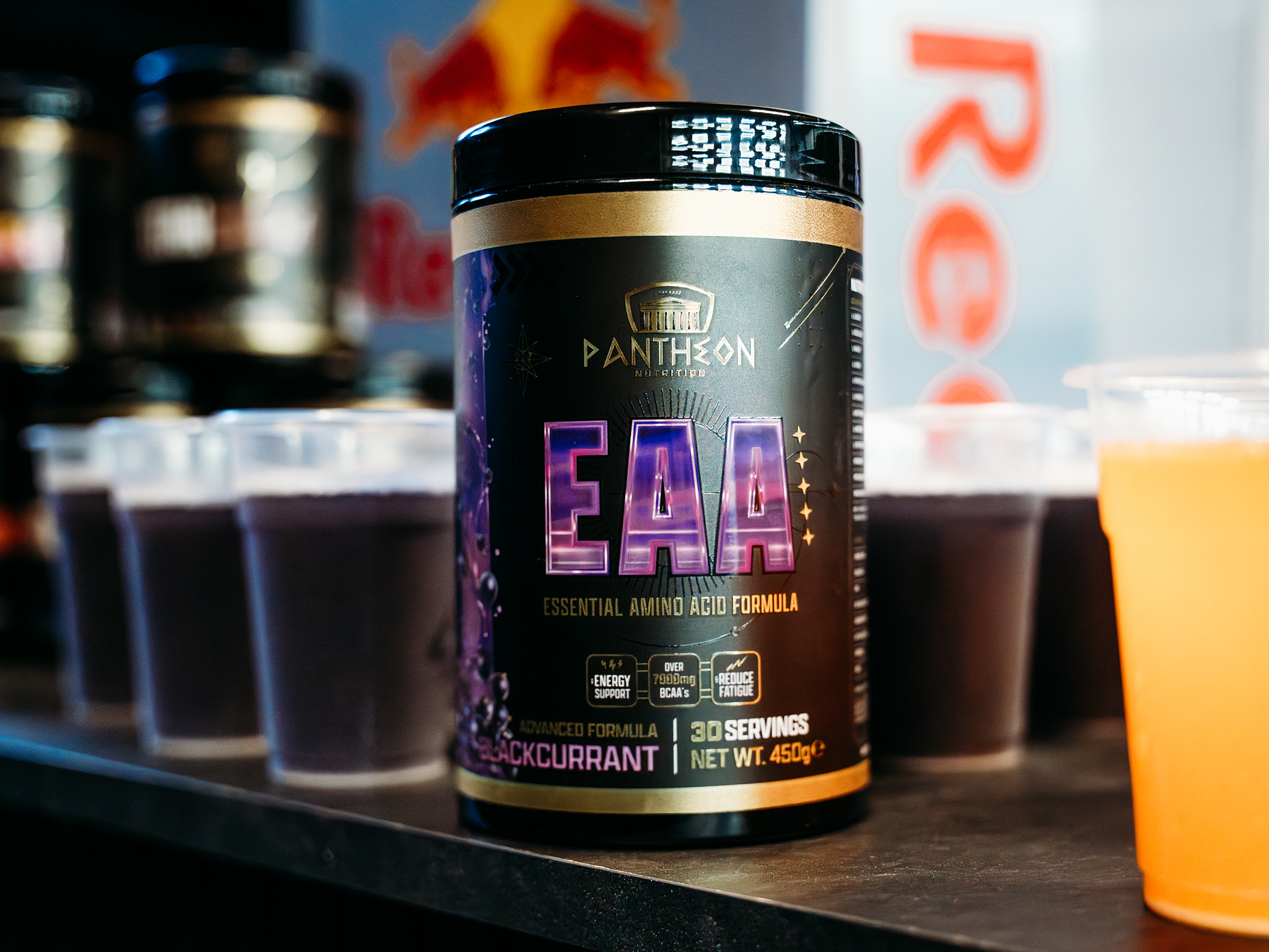 A container of Pantheon Nutrition EAA supplement on a counter surrounded by small sample cups filled with purple and orange drinks, with part of a Red Bull logo visible in the background