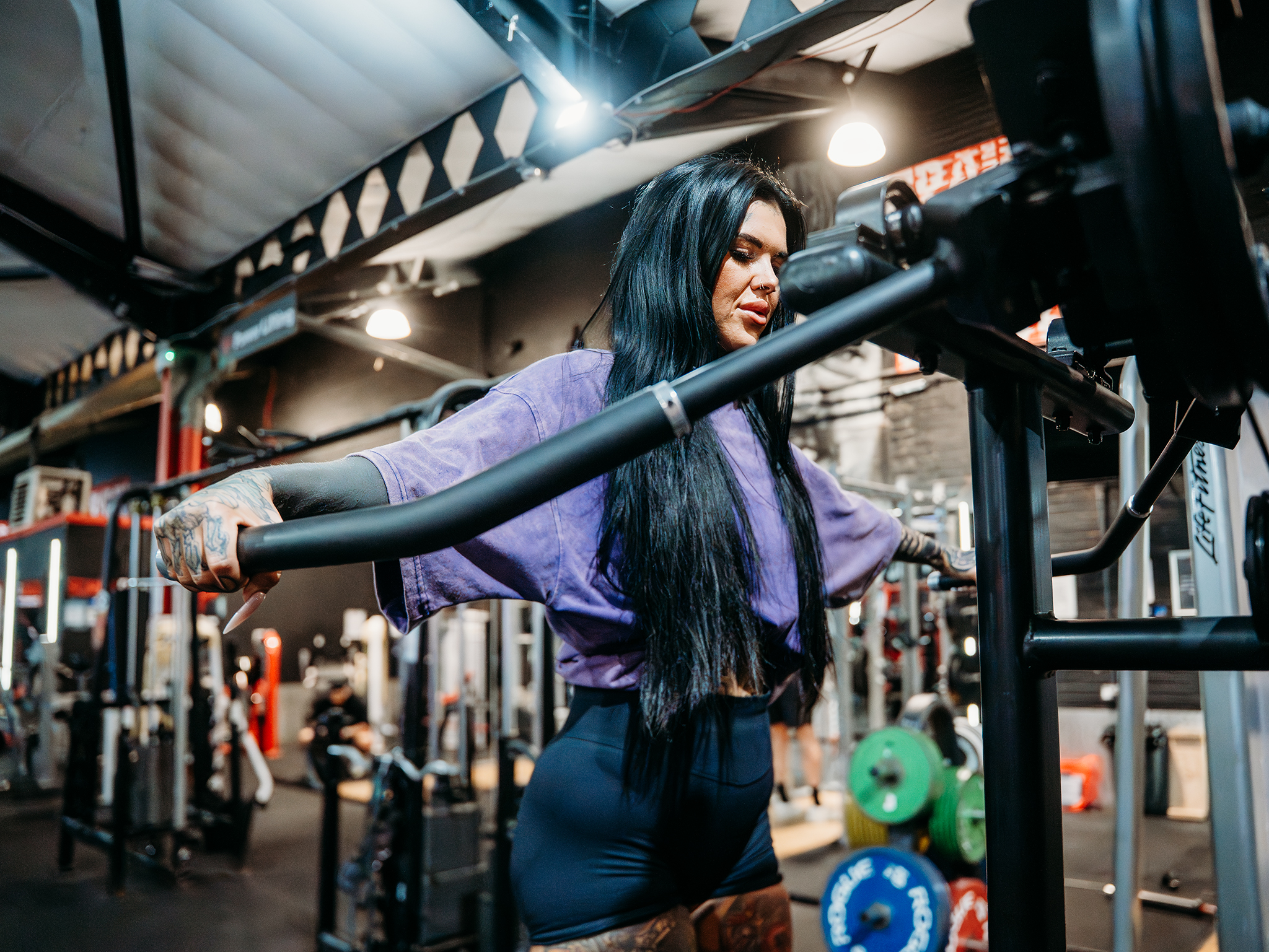 A person stands at a weightlifting machine in a brightly lit gym, wearing a loose purple shirt and dark shorts, with tattoos visible on their arms, surrounded by colorful weight plates and other people working out in the background.