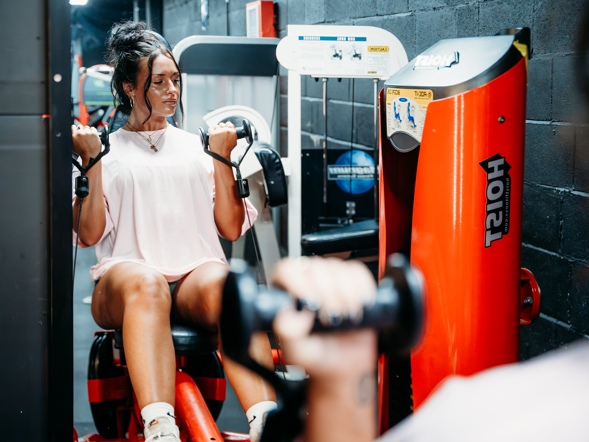 A person uses a red Hoist strength‑training machine to perform a seated bicep curl in a gym, while another person holding a dumbbell is partially visible in the foreground, with additional equipment in the background.