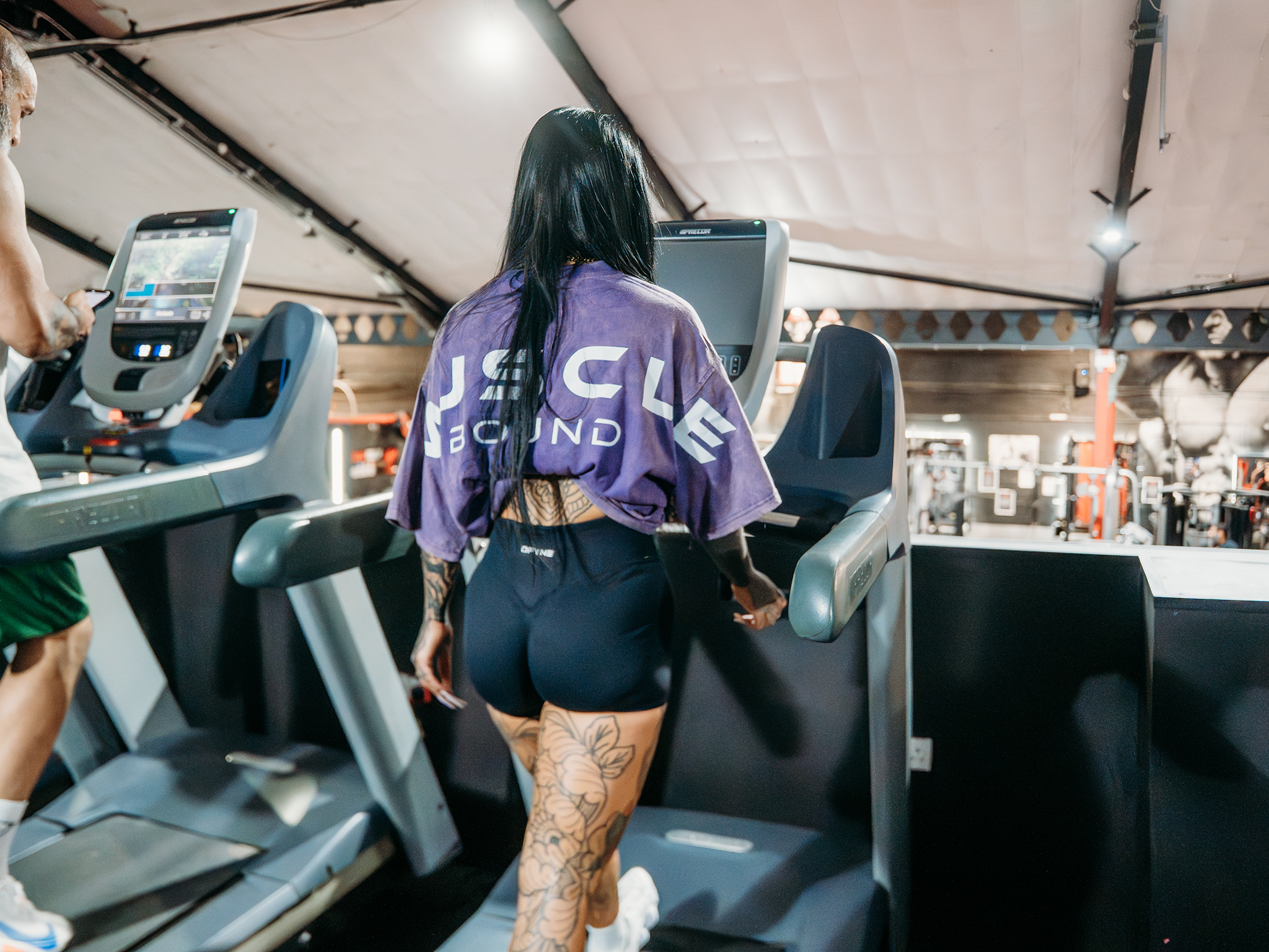 A person walks on a treadmill in a gym, wearing a purple cropped shirt with “Muscle Bound” on the back and black shorts, with tattoos visible on their legs and arm, while another person uses a nearby treadmill in the background.