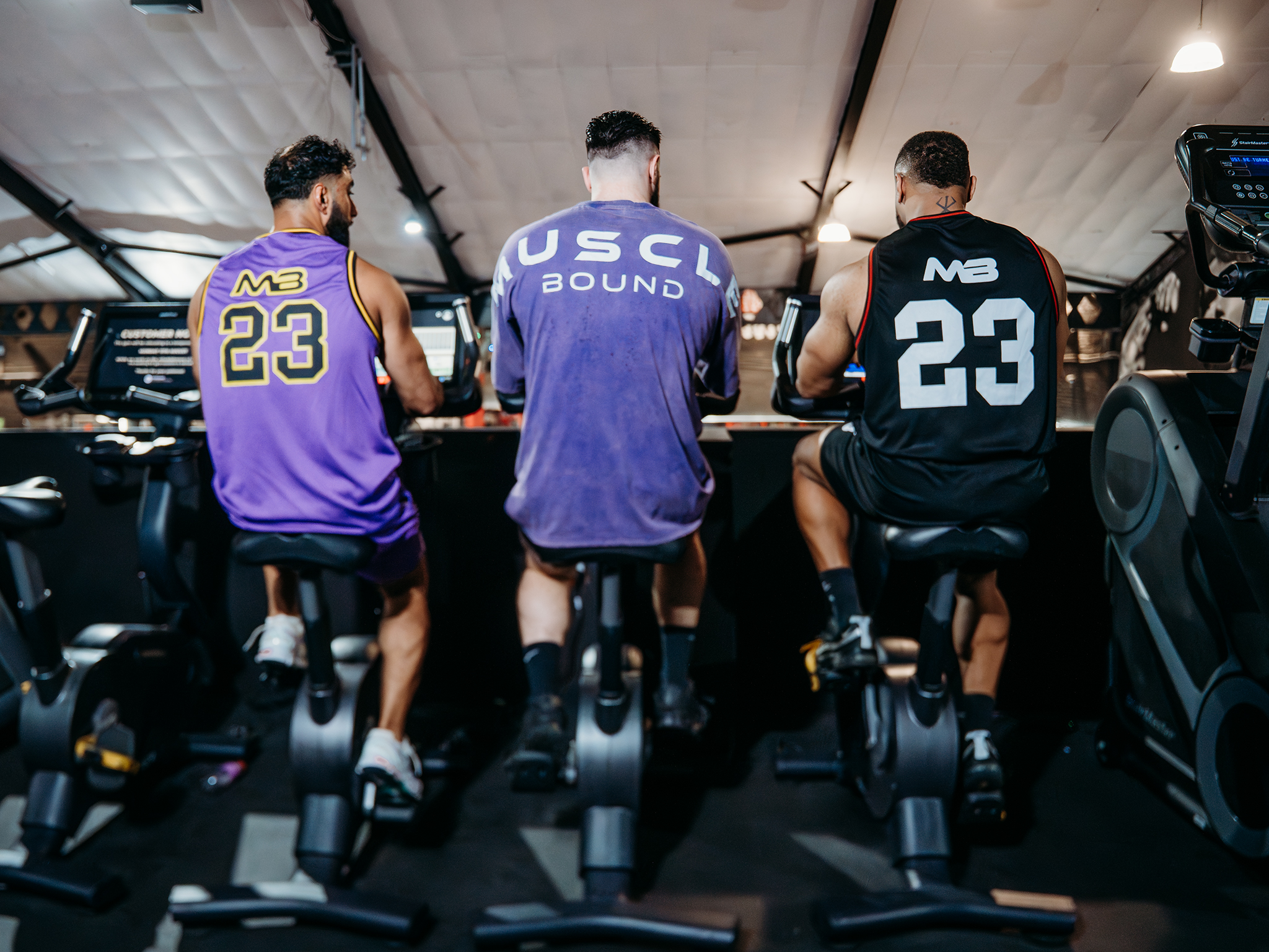 Three people ride stationary bikes in a gym, viewed from behind, wearing purple and black athletic jerseys with numbers on the back, with high ceilings and additional workout equipment visible in the background.