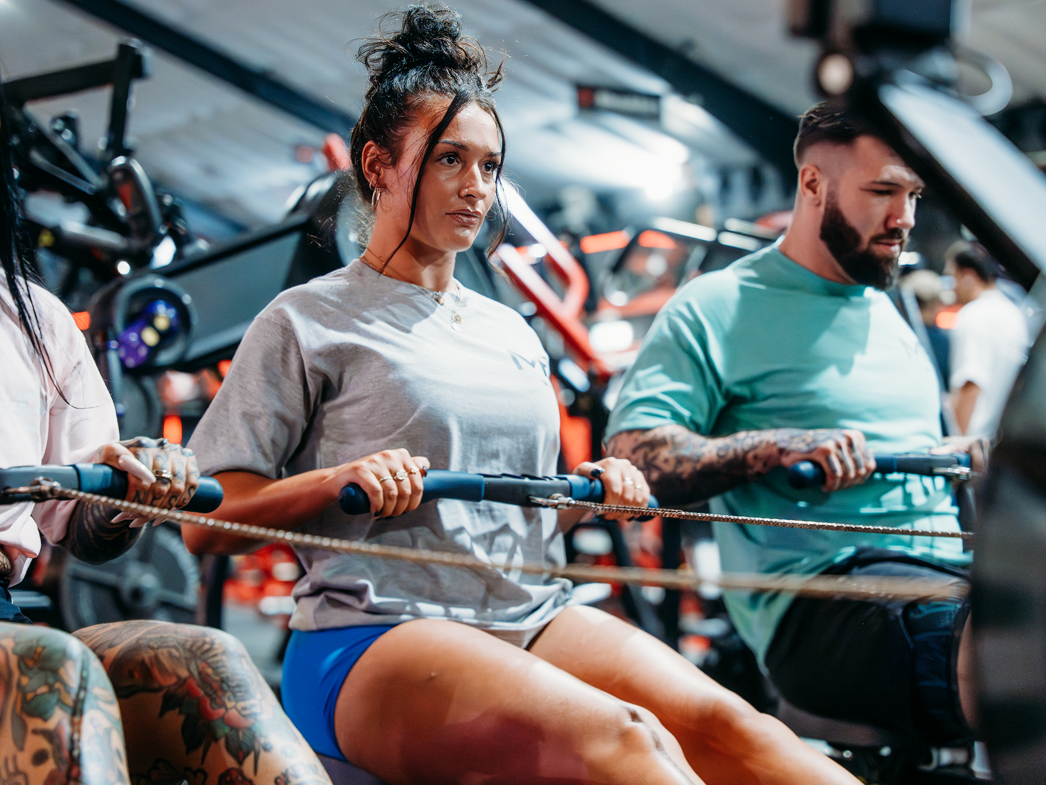 Several people use rowing machines in a brightly lit gym, seated and pulling the handles in unison, with additional strength‑training equipment visible in the background.