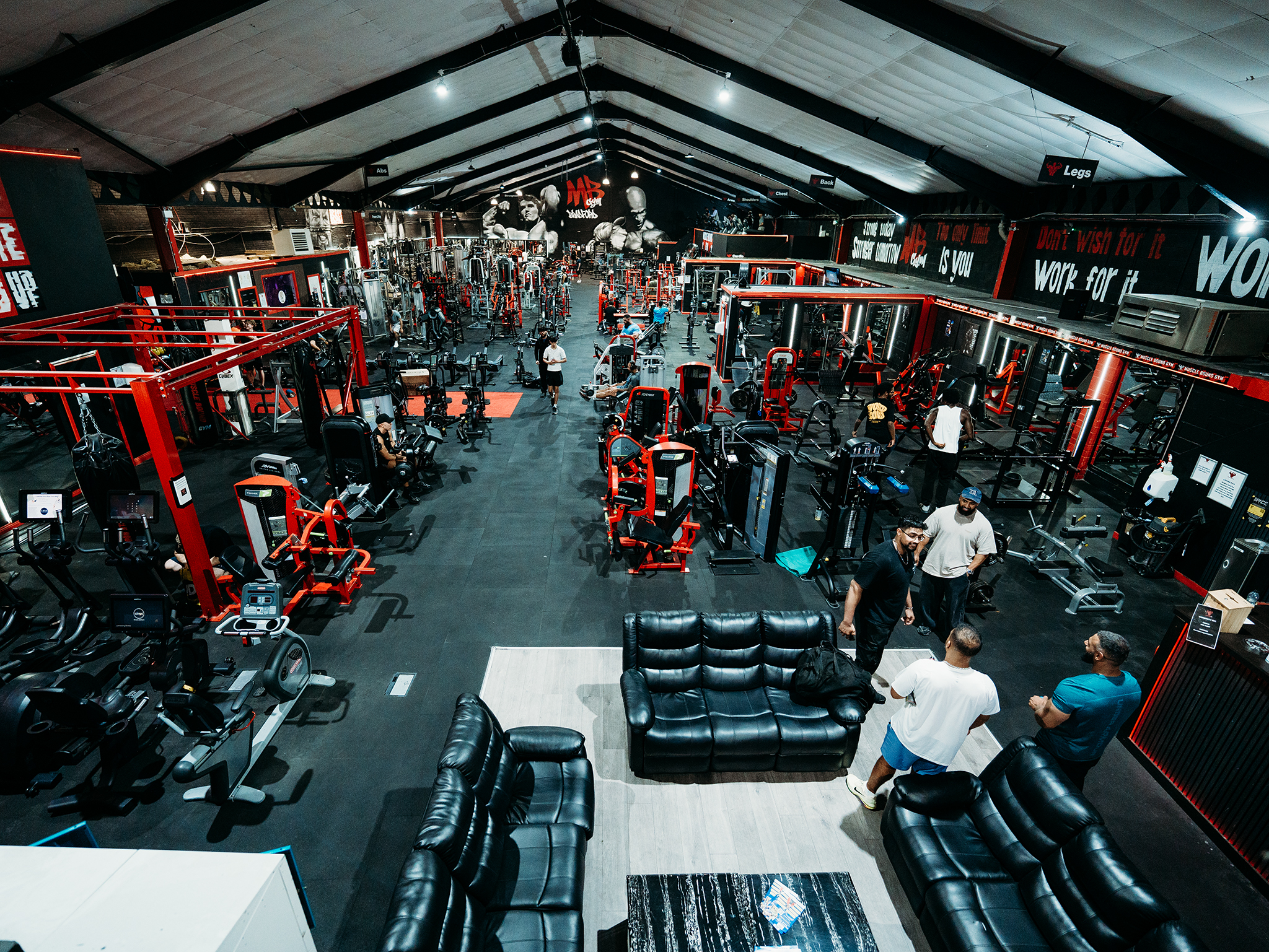 A large, modern gym with high ceilings and bright lighting, filled with red and black exercise machines, with a lounge area of black leather couches in the foreground and people talking nearby, plus motivational quotes on the walls.