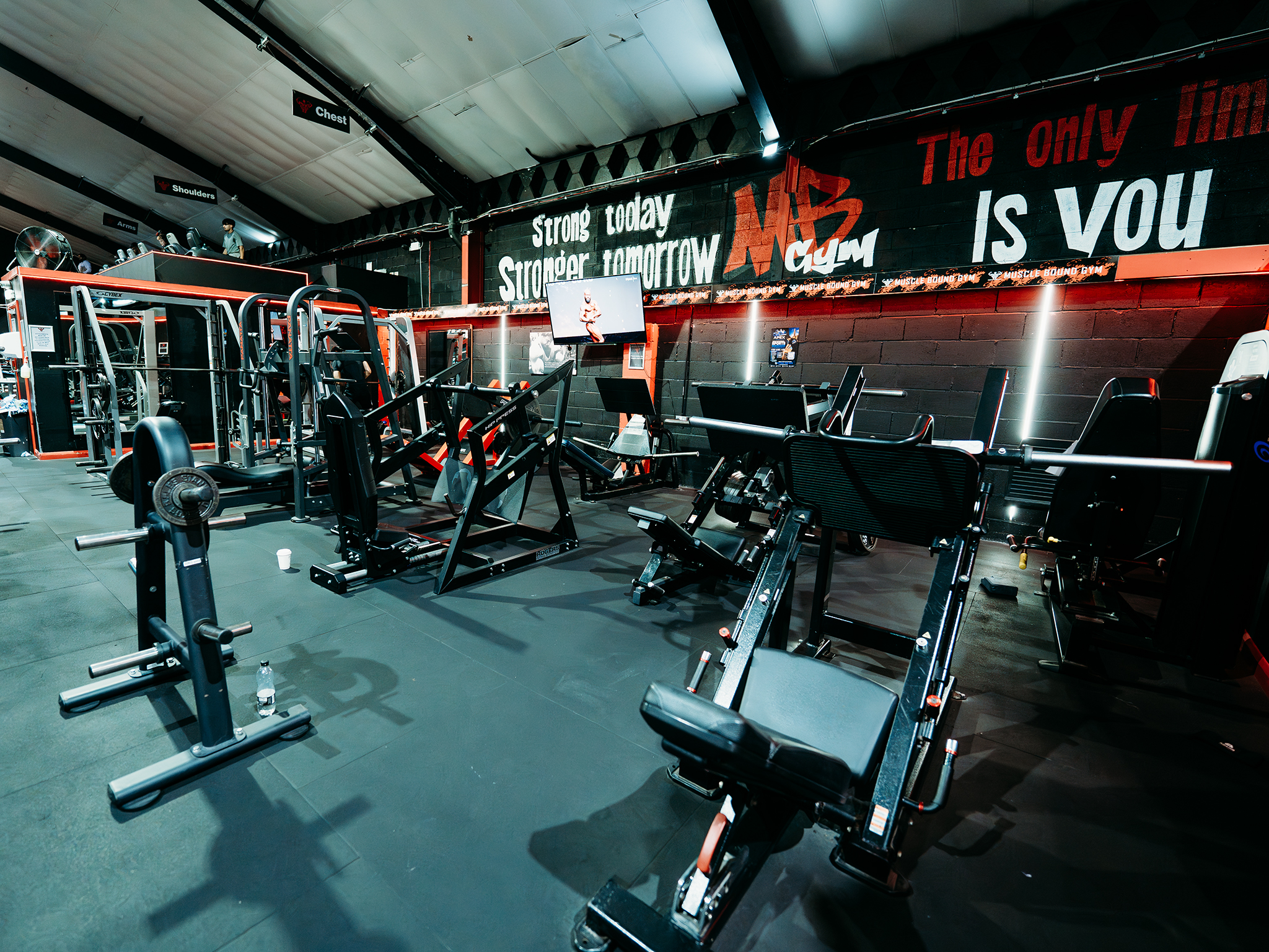 A modern gym with neatly arranged strength‑training machines on a rubber floor, including leg press and squat racks, with overhead signs marking chest, shoulder, and arm zones, and motivational text on the back wall.