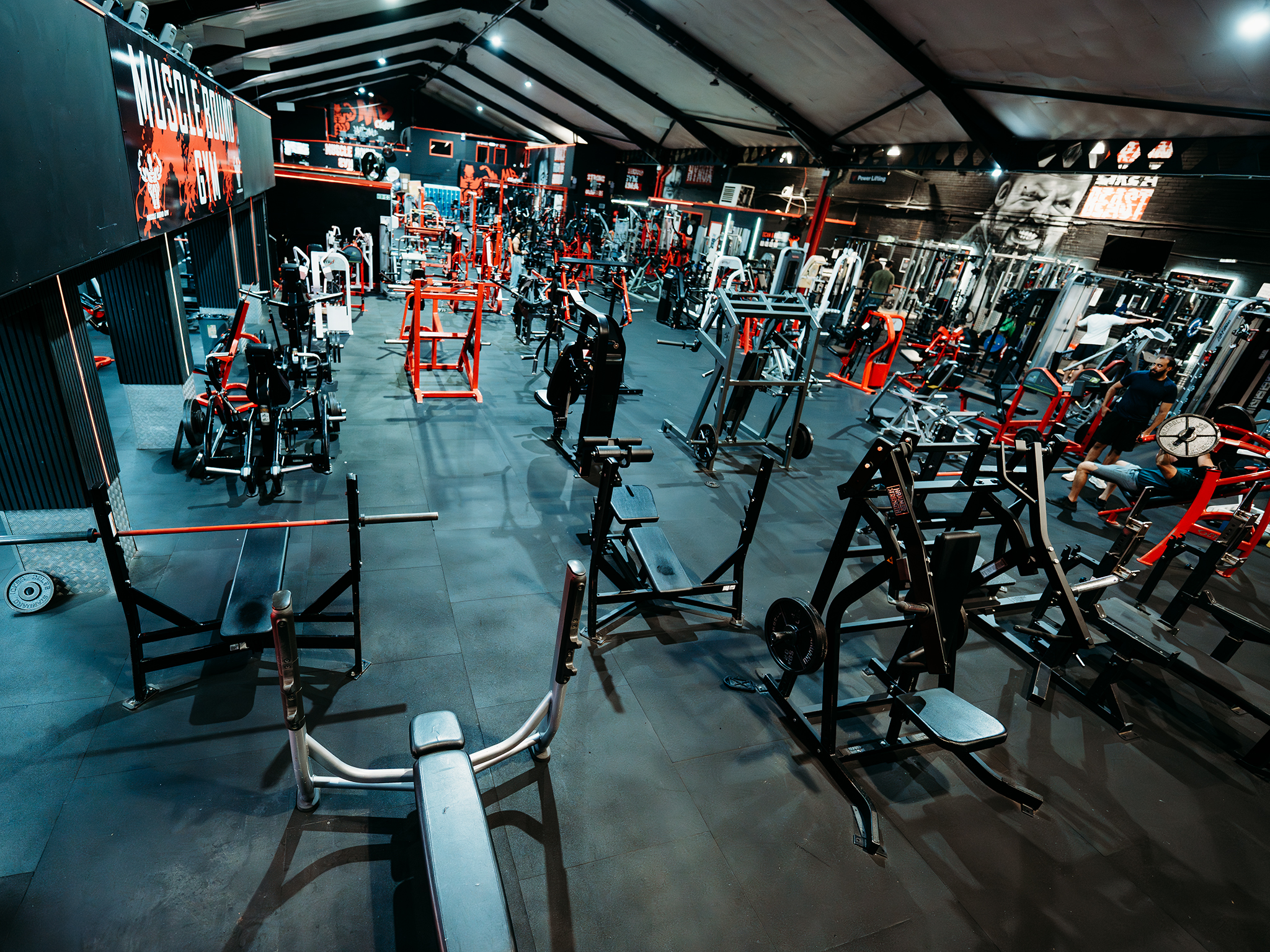 A large, brightly lit gym with high ceilings and exposed beams, filled with red and black strength‑training machines, benches, squat racks, and free weights, with several people working out and motivational signs like “Muscle Building Gym” and “Unleash the Beast” on the walls.
