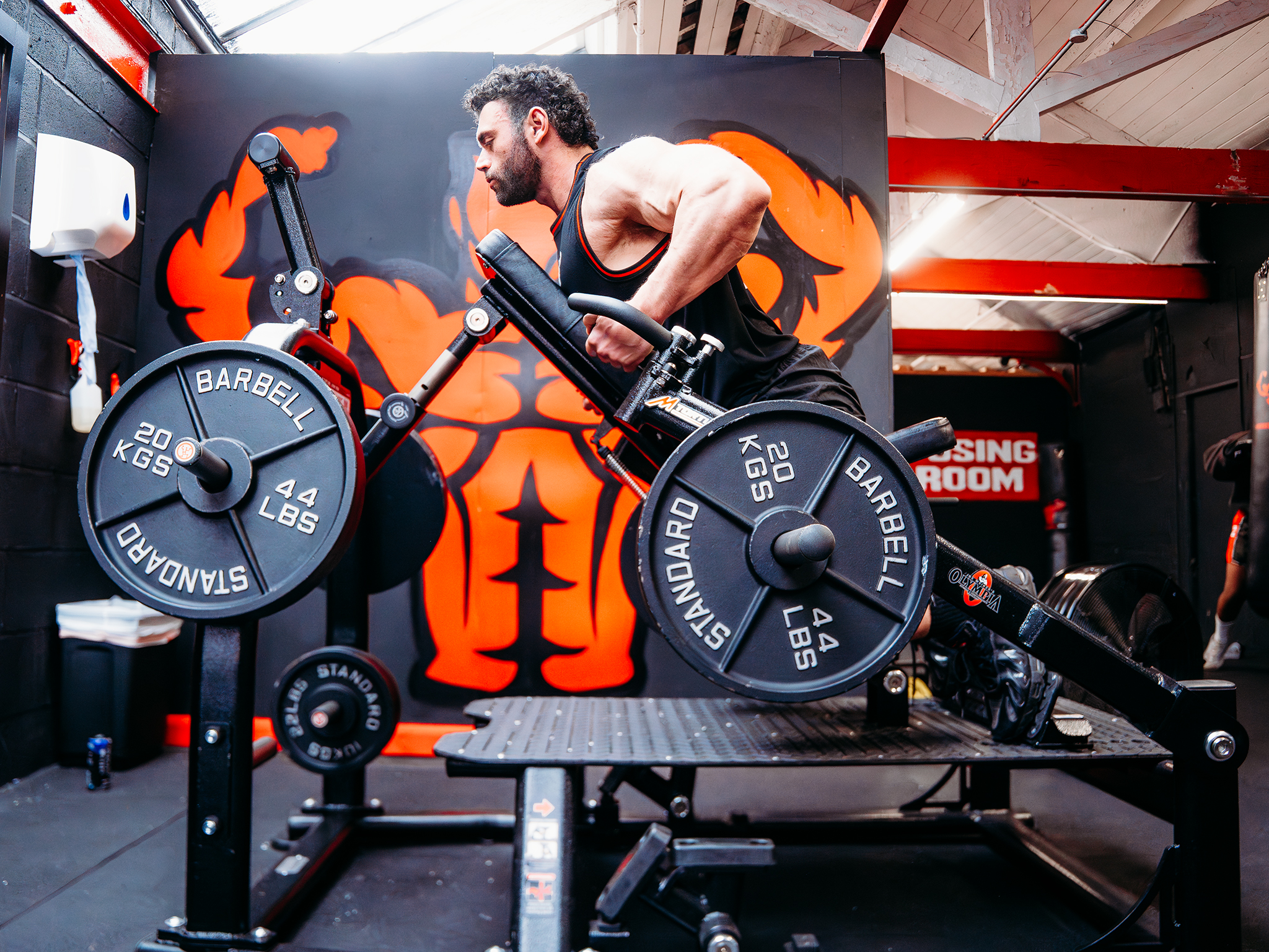 A person performs a chest‑supported row on a gym machine loaded with two 20‑kilogram weight plates. The space has a black‑and‑red color scheme, with a large mural of a muscular figure on the wall and a “DRESSING ROOM” sign visible in the background.
