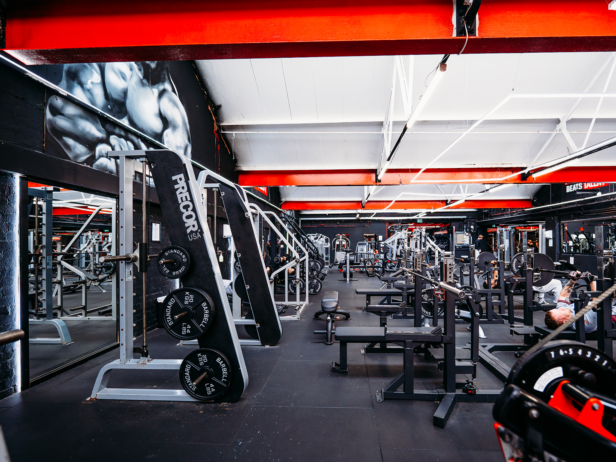 A spacious strength‑training area filled with weight machines, benches, and barbells arranged neatly across the room. Black walls, red beams, and bright overhead lighting create a modern industrial look, with a mural of a muscular figure adding to the gym’s intense, athletic atmosphere.