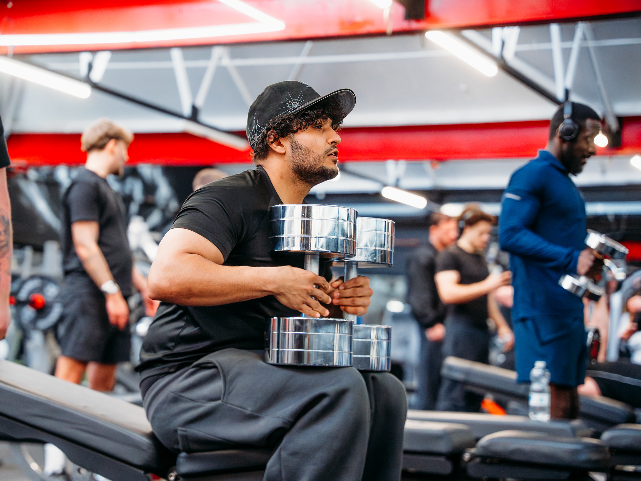 A person seated on a gym bench holding two large chrome dumbbells close to their chest, surrounded by other people lifting weights and various gym equipment under bright lighting and red ceiling beams