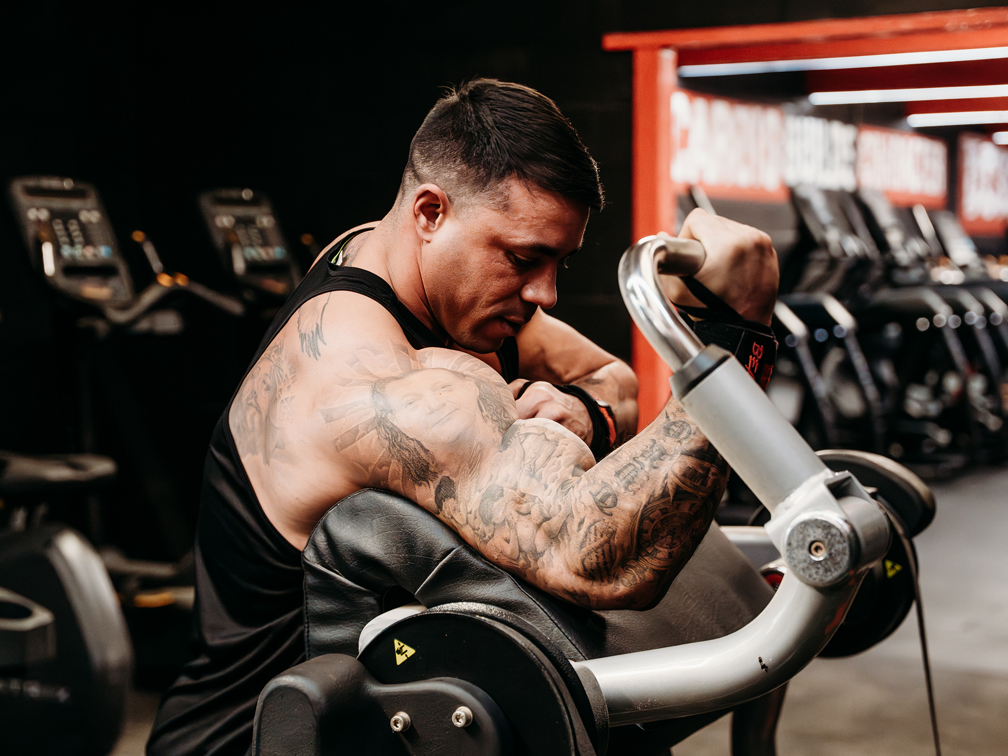 A tattooed person using a preacher curl machine in a gym, gripping the bar and performing a bicep curl while cardio equipment and mirrors appear in the background