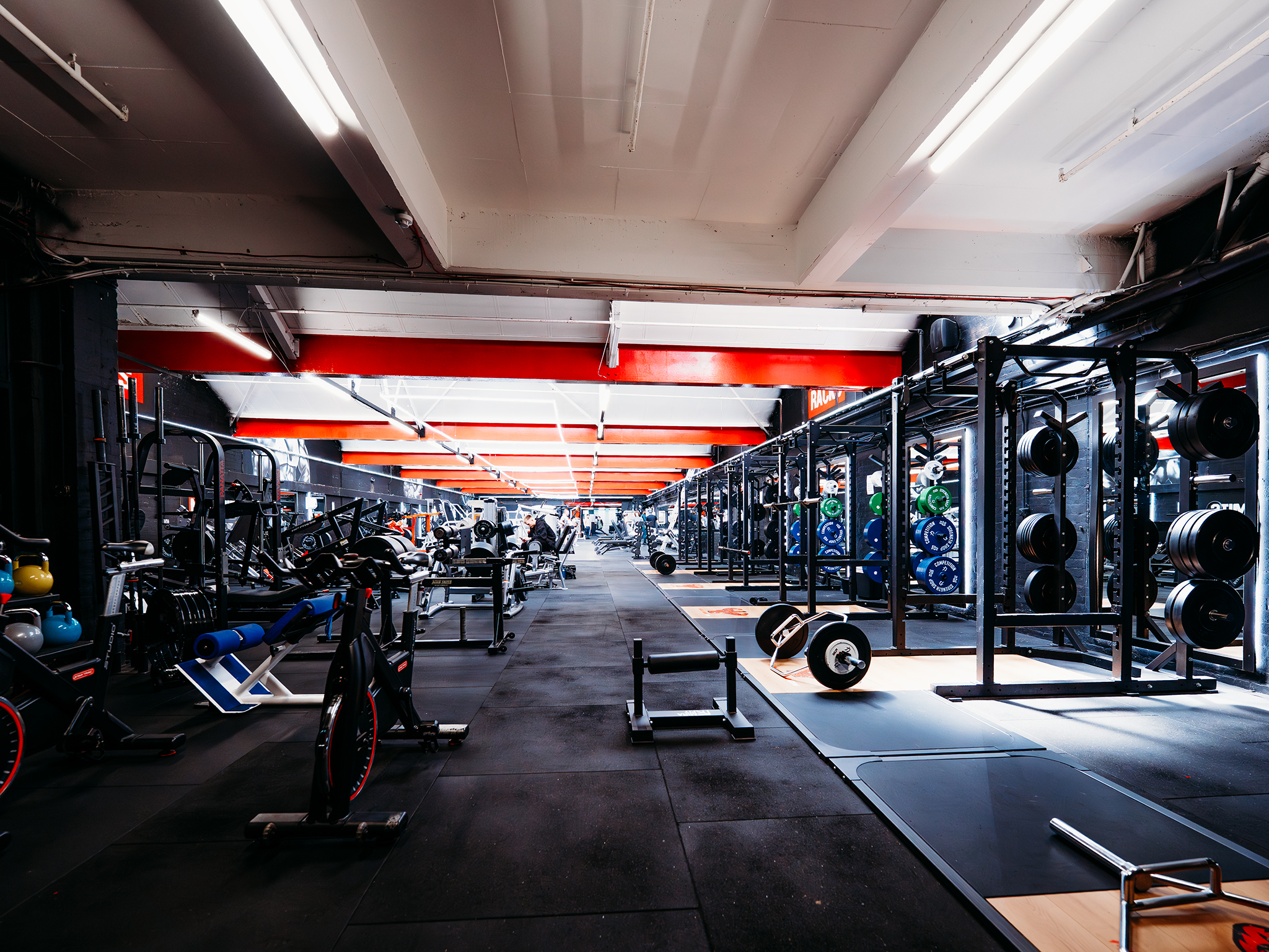 A spacious, modern Musclebound Gym in Huddersfield featuring bright lighting, red ceiling beams, black rubber flooring, and a wide range of strength‑training equipment including squat racks, barbells, kettlebells, and stationary bikes