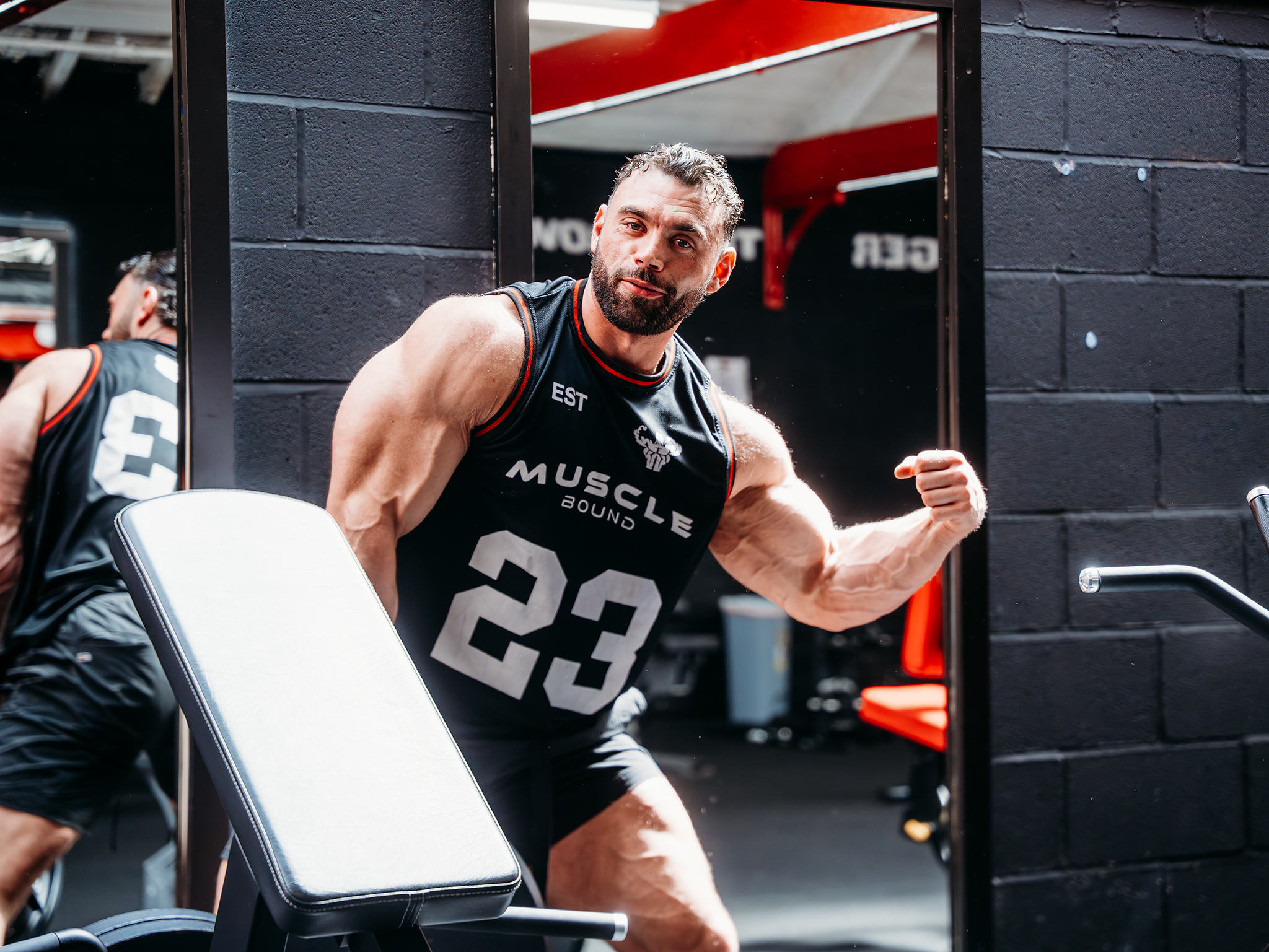 A muscular man posing in front of a gym mirror, wearing a black “Muscle Bound Gym” sleeveless shirt, with red‑and‑black gym equipment visible in the background.