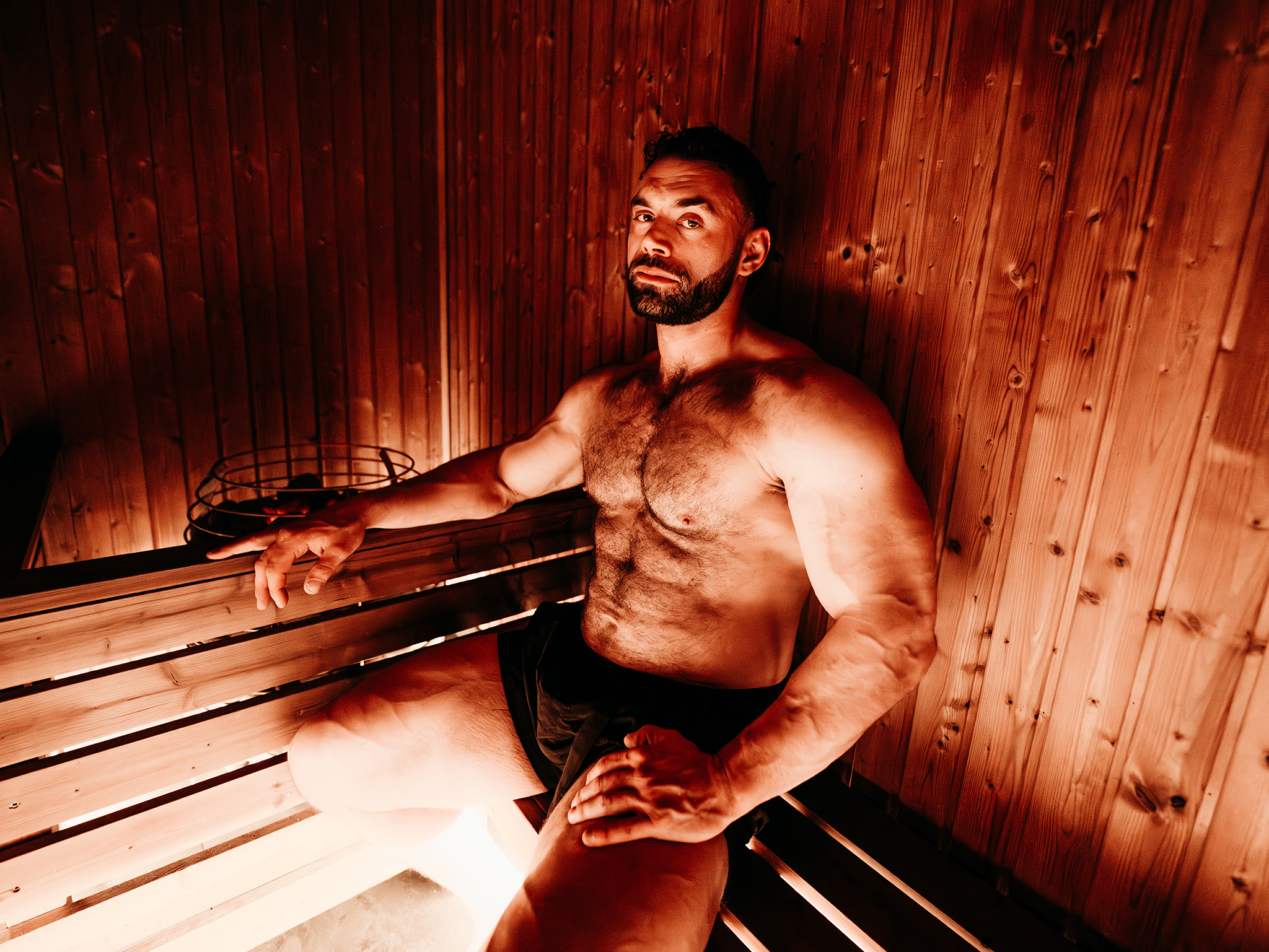 A muscular man sits relaxed inside a wooden sauna, wearing dark shorts and resting one arm on the bench. Warm, reddish lighting highlights the wood panels and a metal basket of sauna stones nearby.