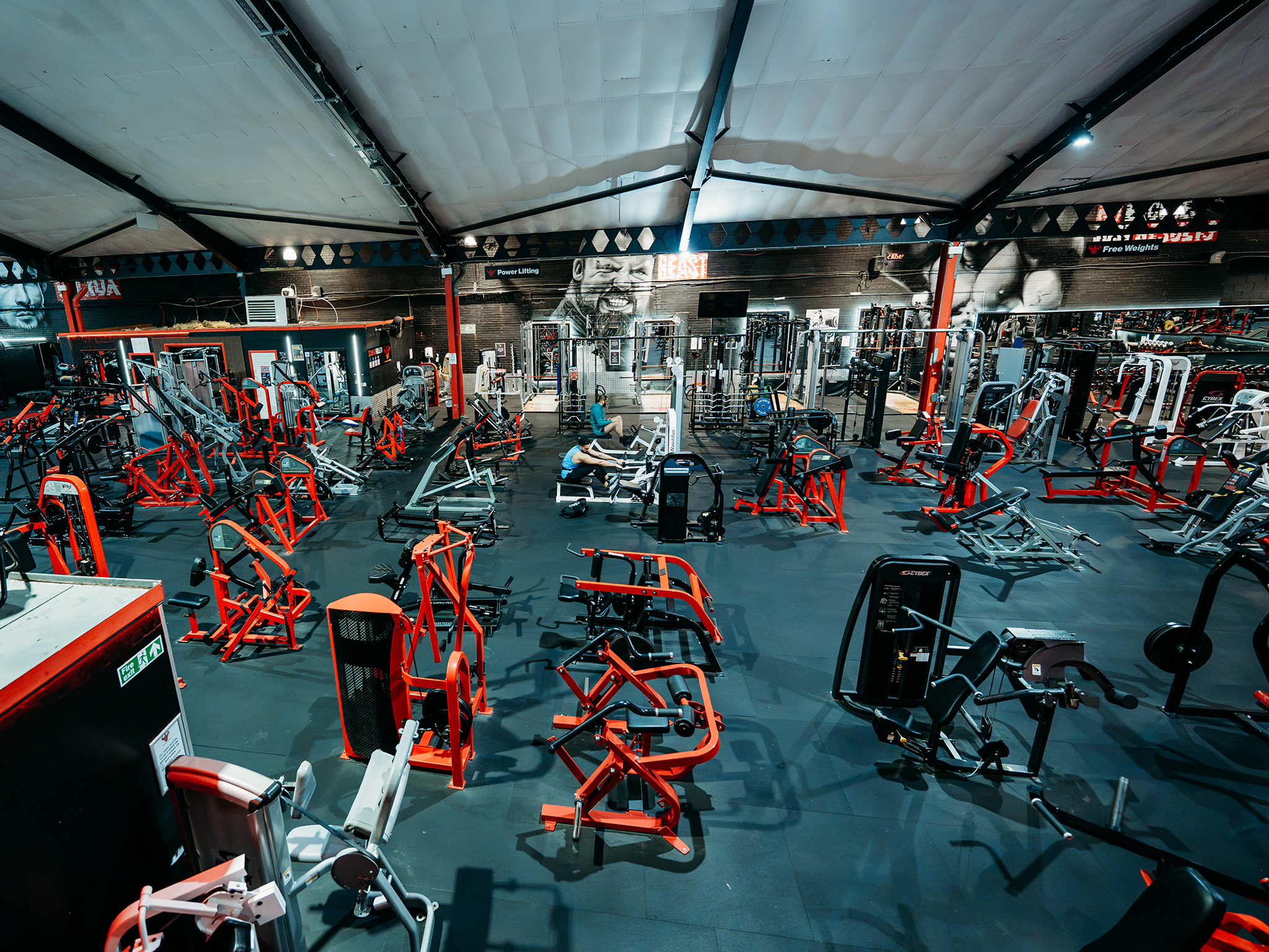 Spacious strength-training gym featuring a wide range of resistance machines and free-weight equipment, with black flooring, red-accented machines, and high industrial-style ceilings.