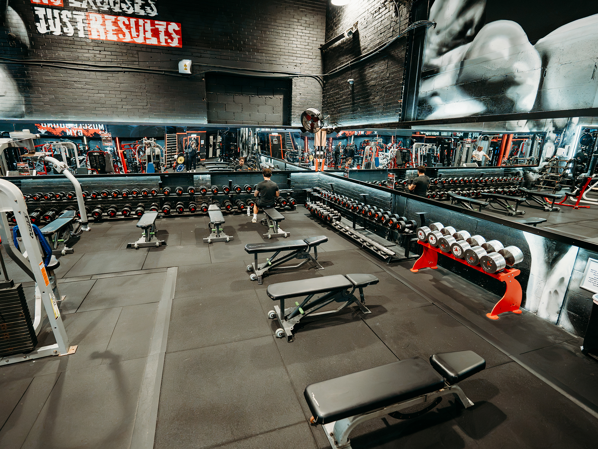 Free-weights section with multiple adjustable benches, extensive dumbbell racks, mirrored walls, and motivational gym graphics in a modern fitness facility.