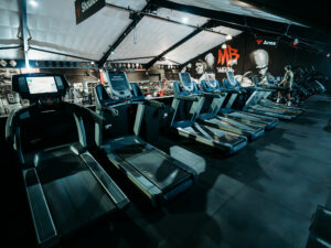 Modern cardio training area with a row of treadmills and exercise machines, set within a spacious gym featuring industrial décor and fitness-themed wall murals.