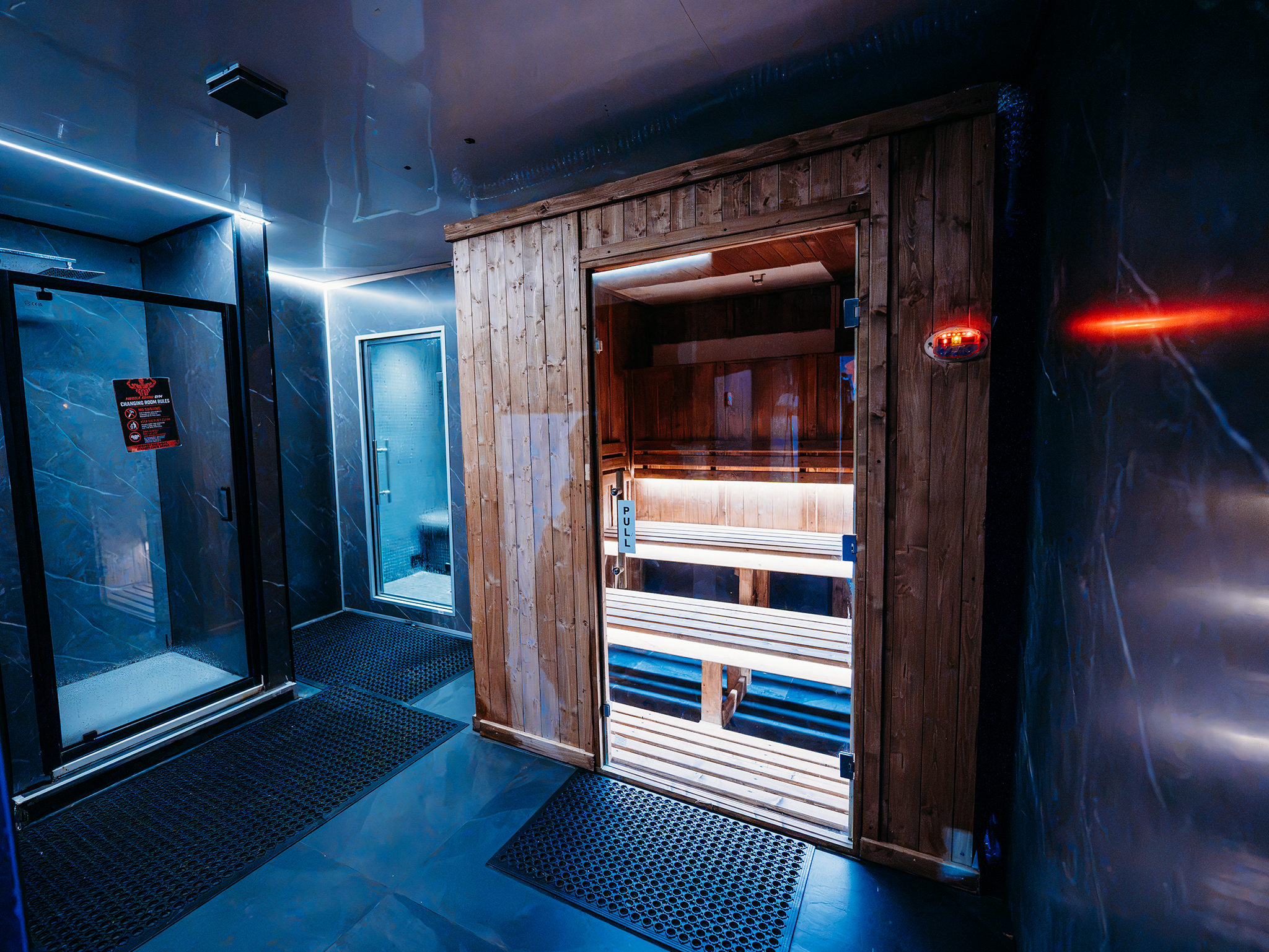 Contemporary spa room with a wooden sauna cabin, glass door, dark stone-effect walls, LED accent lighting, and rubber safety flooring.