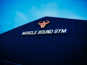 Exterior of Muscle Bound Gym with illuminated signage mounted on a dark metal-clad building against a blue evening sky.