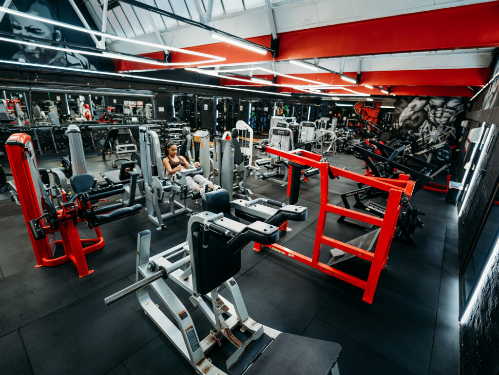 Large gym floor equipped with multiple strength-training stations, selectorised machines, and free-weight equipment. Bright linear lights illuminate the space, which has black walls and bold red structural beams. A person is using a leg machine near the centre, while a red-framed machine stands prominently in the foreground. Mirrors line the walls, reflecting the equipment throughout the room.