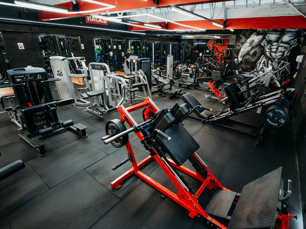 Interior of a strength-training gym featuring a variety of resistance machines arranged across a spacious workout area. Black flooring contrasts with red ceiling beams and red accents on equipment. A seated exercise bench occupies the foreground, while additional machines, mirrors, and a person using equipment are visible in the background.