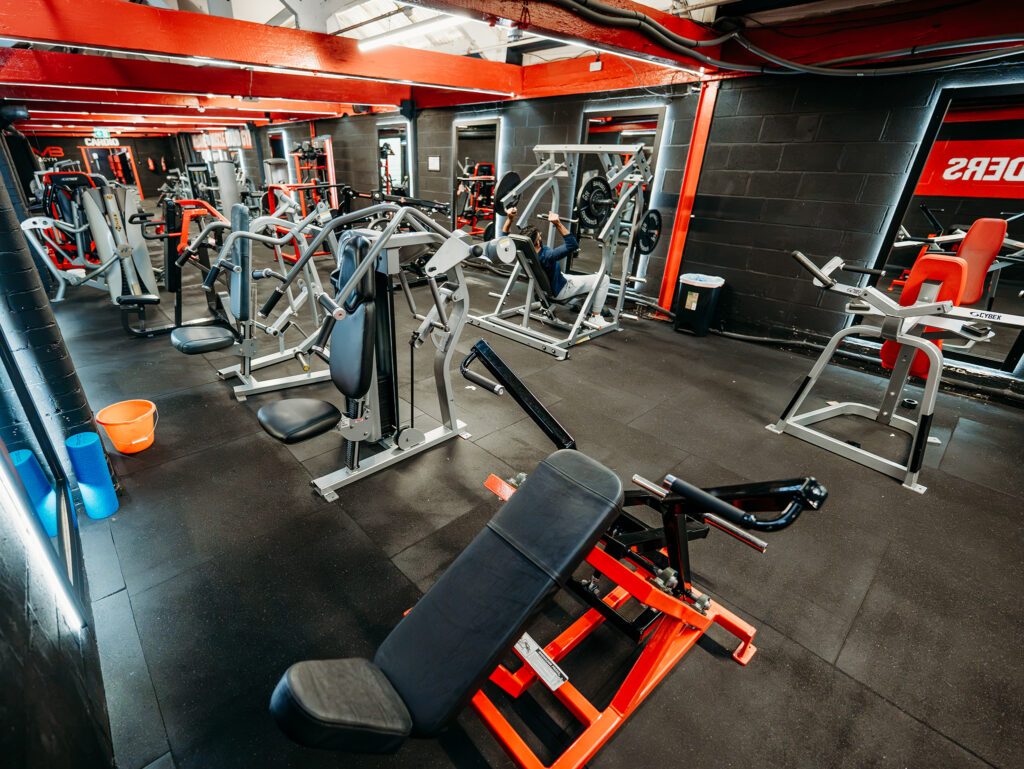 Wide view of a modern gym with black walls, red ceiling beams, and bright overhead strip lighting. Numerous strength-training machines and squat racks fill the room. A red-and-black 45-degree leg press machine is prominently positioned in the foreground. Weight plates are stored along the back wall, and a large monochrome mural of a muscular figure is painted on the right wall.